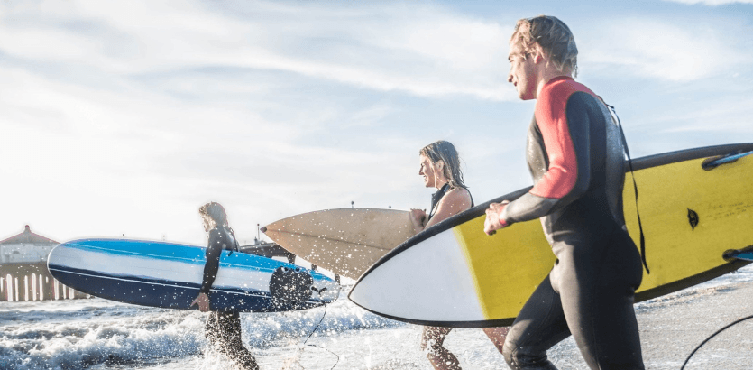 Three individuals in wetsuits are carrying surfboards, preparing to head into the ocean for a surfing session.