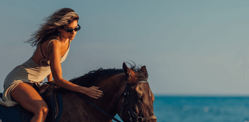 A woman elegantly rides a horse along the shoreline, with waves gently lapping at the beach in the background.