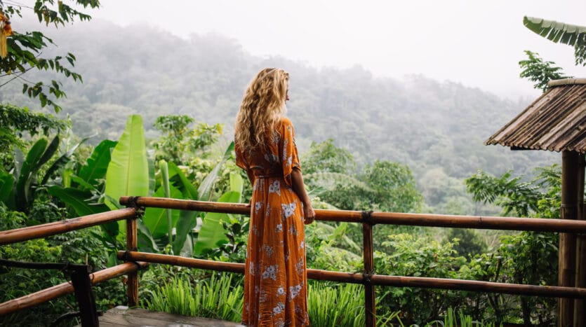 Back view of unrecognizable female tourist with long hair standing near bamboo fence on wet wooden plank of guest house in tropical garden with green plants and trees