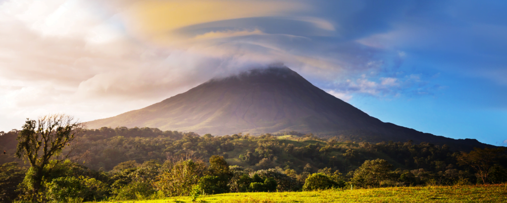 A large volcano rises in the distance, surrounded by clouds in a clear blue sky.