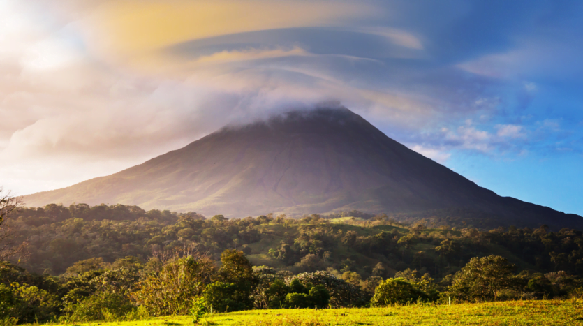 A large volcano rises in the distance, surrounded by clouds in a clear blue sky.