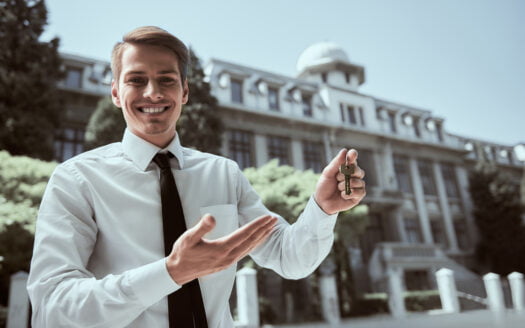 Successful realtor in white shirt showing keys and smiling at camera.