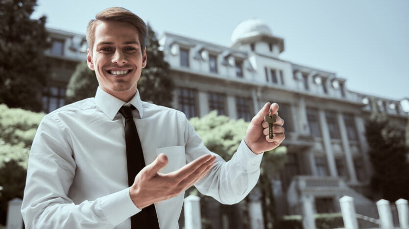 Successful realtor in white shirt showing keys and smiling at camera.